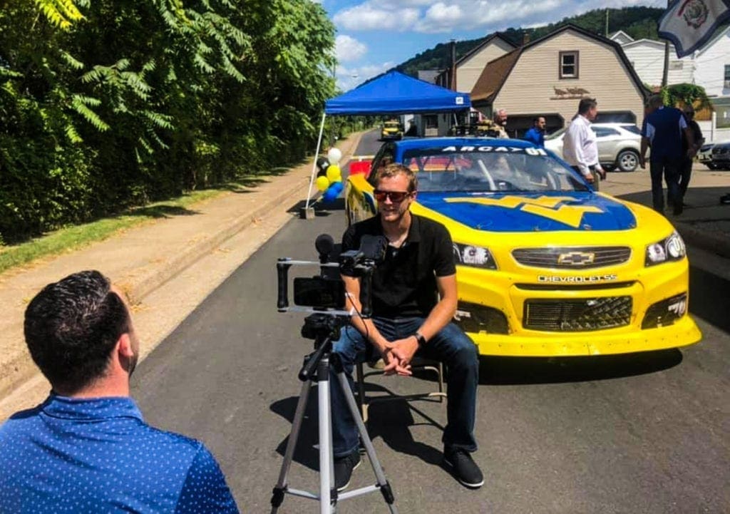 A race car driver being interviewed while his WVU stock car resting behind him.