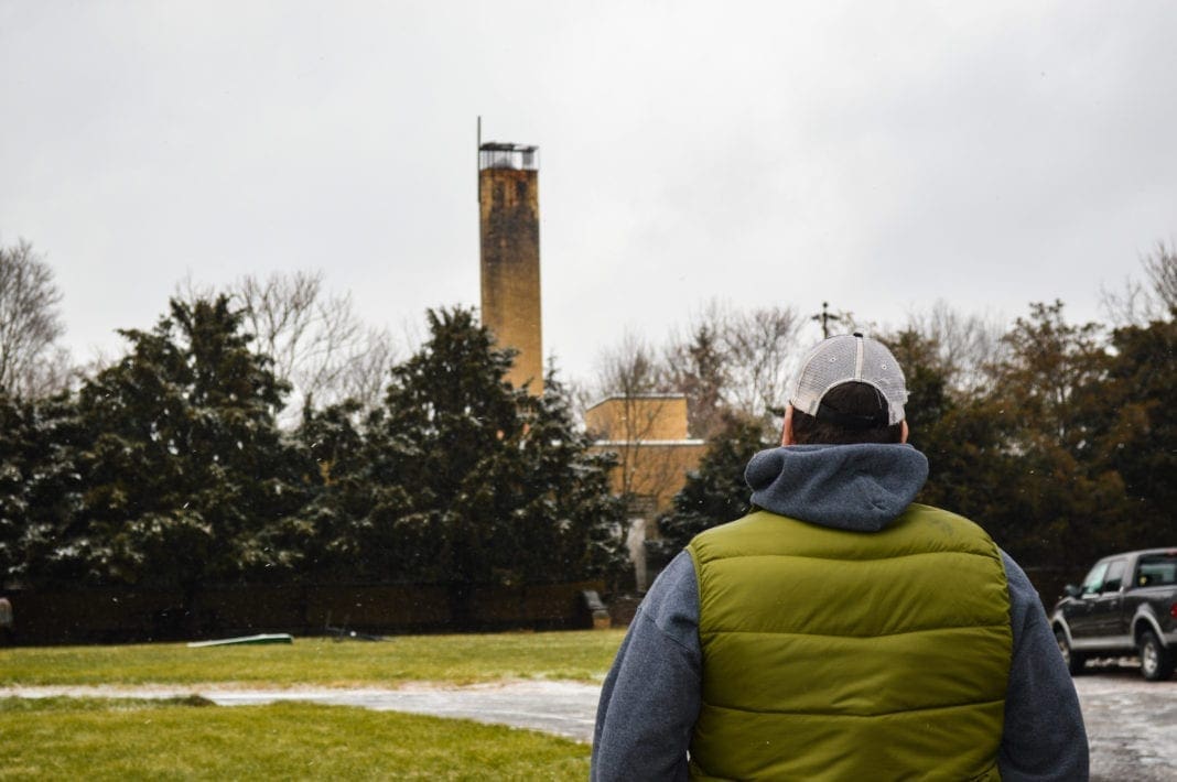 A photo of a man standing in from of a one story hospital that has a high smoke stack.