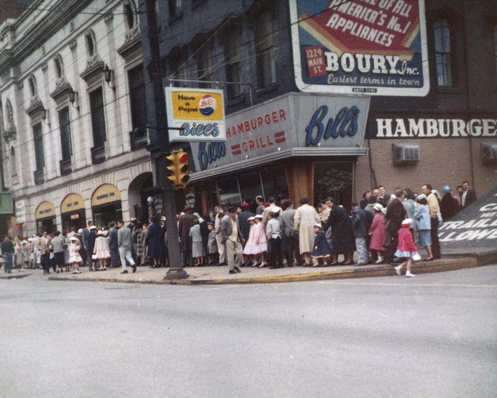 A long line waiting to get into a theatre. in front of Bill’s Hamburger Grill