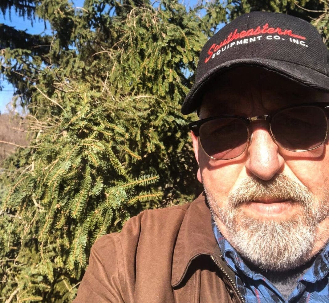 A phone of a man in front of a pine tree.